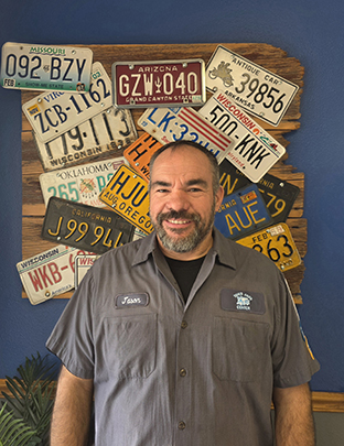 Tom's Auto Center - Jason, ASE World Class Technician, standing in front of wall of license plates